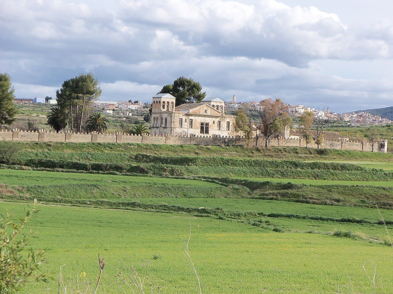 Campos verdes en bancales y, al fondo, un caserón con torre tras una tapia; detrás, las casas de Bullas bajo un cielo de nubes.